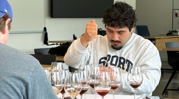 Undergraduate student measures wine in a pipette with several glasses of red wine on a table in front of him.