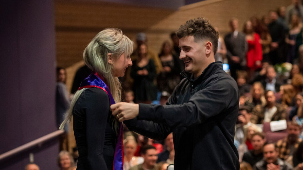 Friend/family member puts stole with pin around neck of graduating nurse at December Pinning Ceremony in front of a crowd