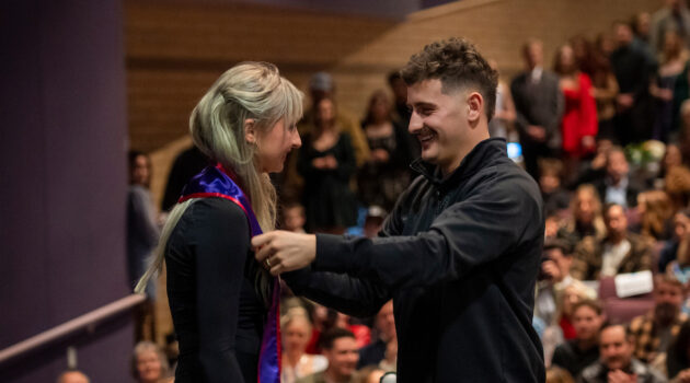 Friend/family member puts stole with pin around neck of graduating nurse at December Pinning Ceremony in front of a crowd