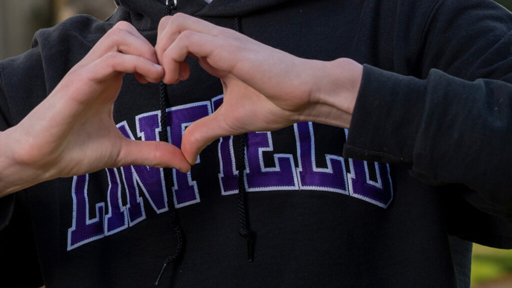 Close up of hands in a heart shape in front of a black sweatshirt, with purple text "Linfield"