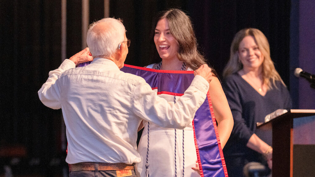 An older man puts the stole over a graduating female nursing student. Faculty member Jess Nesher at podium in background.