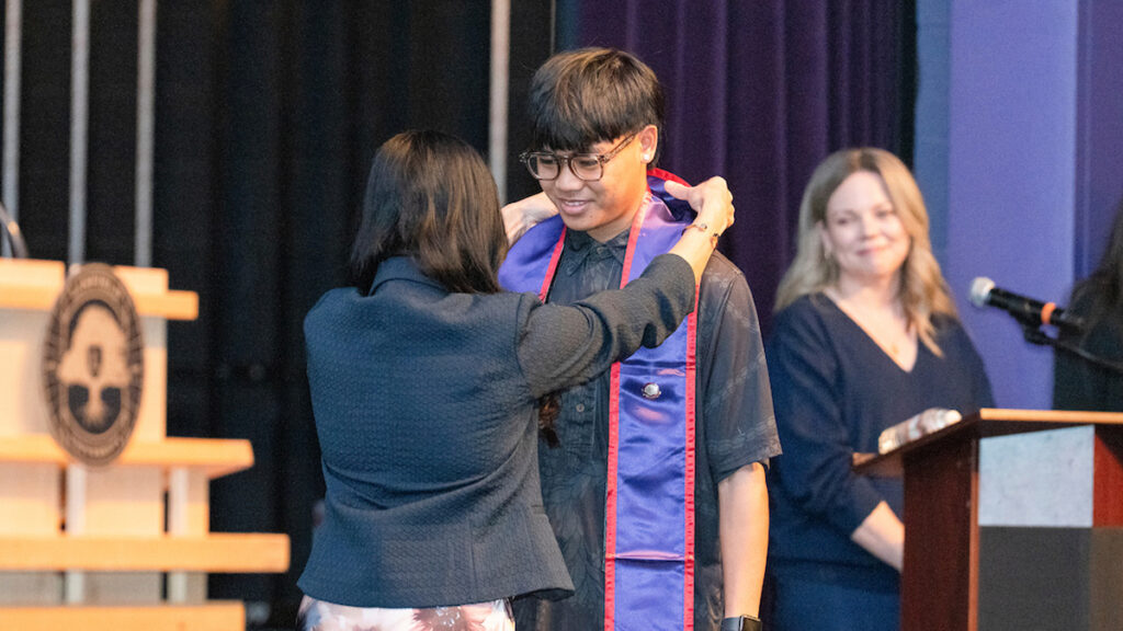 Male nursing student receives stole from a female guest. Jess Nesher at podium in back.