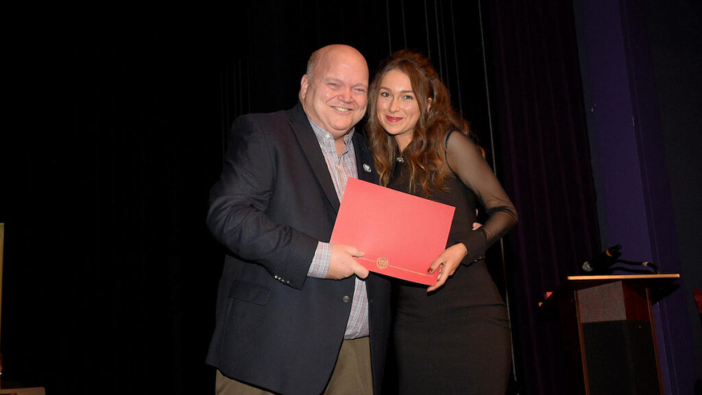 Dean Paul Smith stands with graduating student Lauren Holford on stage, each holding a corner of a red certificate folder.