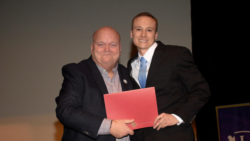 Dean Paul Smith stands with graduating student John Calder on stage, each holding a corner of a red certificate folder.