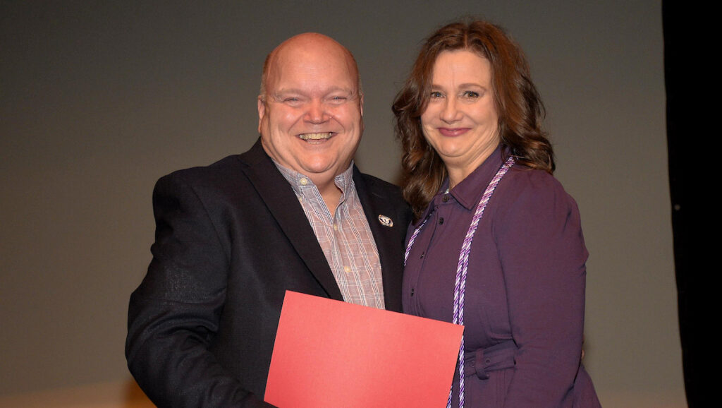 Dean Paul Smith stands with graduating student Julie Peterson on stage, each holding a corner of a red certificate folder.