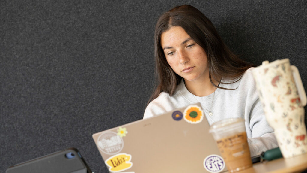 female student sits behind a laptop, covered with floral and LInfield stickers. Next to her is an iced coffee and a Stanley-style cup.