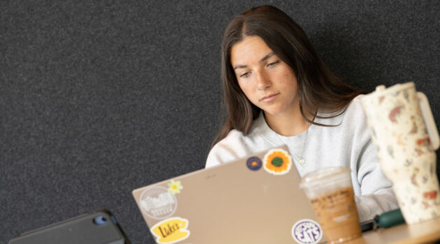 female student sits behind a laptop, covered with floral and LInfield stickers. Next to her is an iced coffee and a Stanley-style cup.