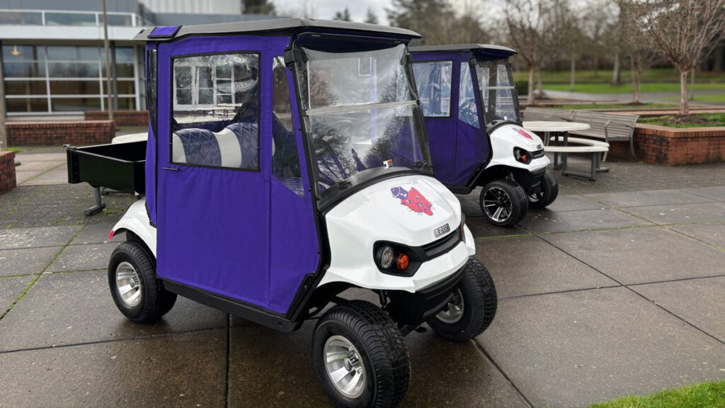 Two electric golf-cart style vehicles parked on the Linfield Portland campus, with Linfield logos on them and purple weather doors.
