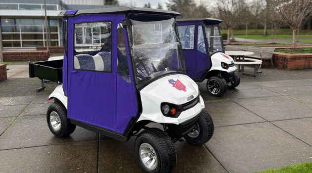 Two electric golf-cart style vehicles parked on the Linfield Portland campus, with Linfield logos on them and purple weather doors.