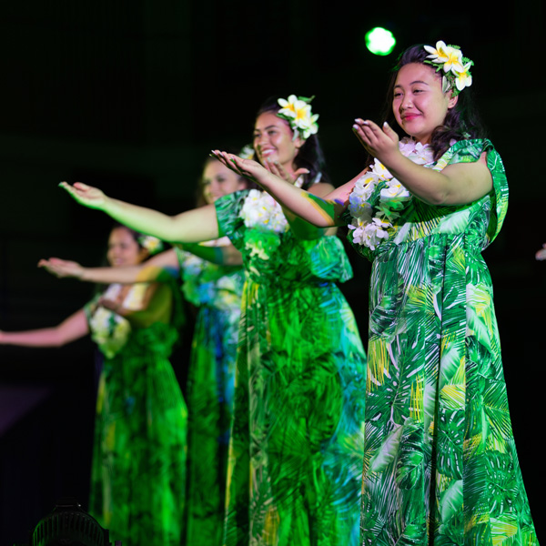 Diagonal line of four women in green palm leaf print dresses and plumeria in their hair perform dance