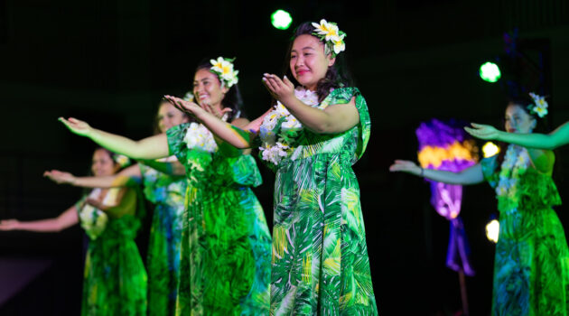 Diagonal line of four women in green palm leaf print dresses and plumeria in their hair perform dance