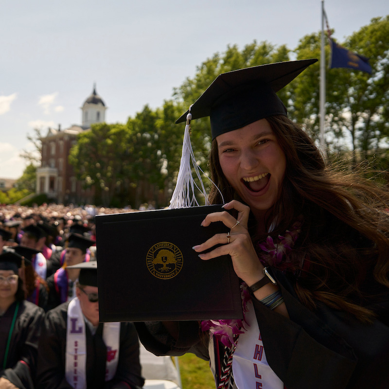 Graduate in cap and gown poses for camera with diploma and a big smile with the crowd of graduates and Pioneer Hall behind her.