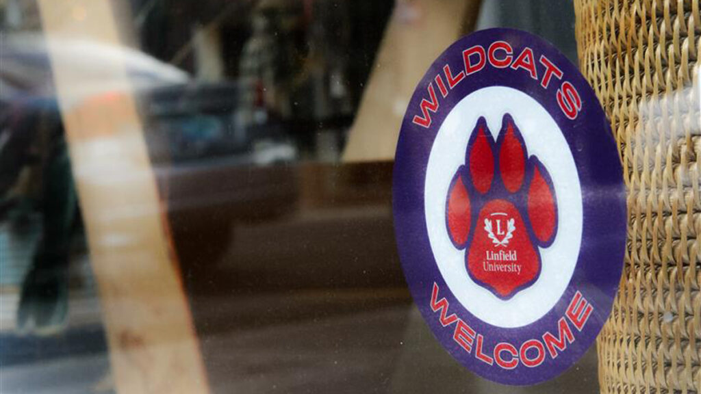 A Cats Community Partner sticker is displayed in a business window.The sticker is a purple circle reading Wildcats Welcome. The interior of the sticker has a red and purple paw on a white background with the Linfield logo.