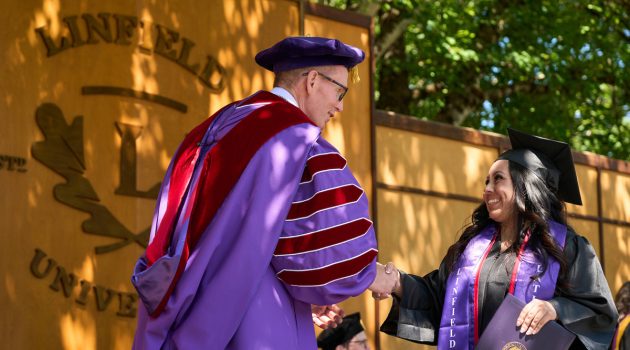 At Commencement 2026, President Blegen shakes the hand of a graduate who is holding her diploma. Linfield University logo on stage in background.