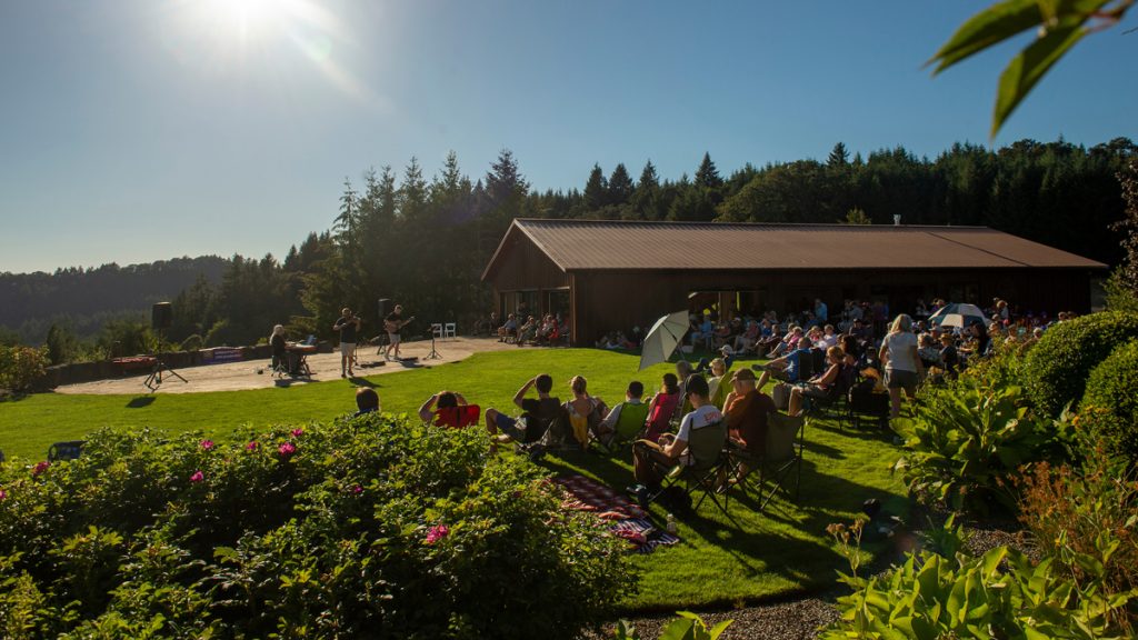 Group of people sit in chairs outside around an outdoor concert stage. Event building on the right.