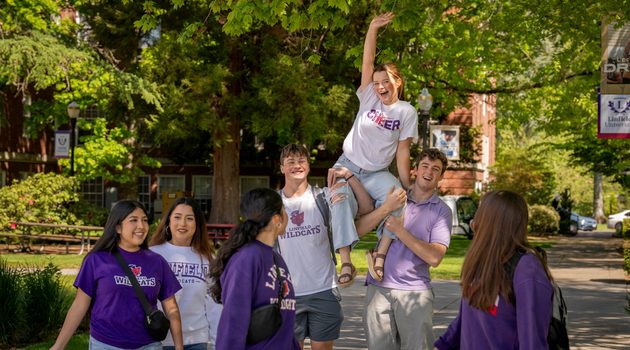 A group of students walk on campus. Two are holding up one on their shoulders.